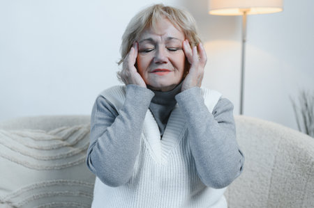 Senior woman sitting on a sofa at home with a headache, feeling pain and with an expression of being unwell.の写真素材