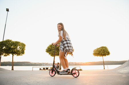 Child girl goes down a ramp with a scooter in a skate park on a sunny summer day.