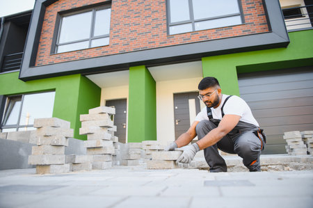 Indian master in gloves lays paving stones in layers. Garden brick pathway paving by professional paver worker. Laying gray concrete paving slabs in house courtyard on sand foundation base.の写真素材