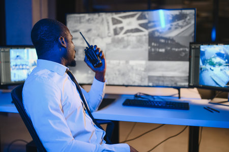African American security guard looking at desktop computer screen while speaking on walkie talkie.の写真素材