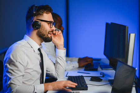 Male Data Scientist Works on Personal Computer Wearing a Headset in Big Infrastructure Control and Monitoring Room. Young Engineer in a Business Call Center Office Roomの写真素材