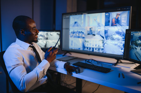 African American security guard looking at desktop computer screen while speaking on walkie talkie.