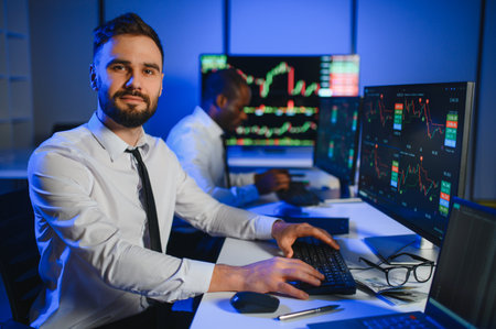 Financial Analyst Talking to Investment Banker in the Evening at Work. Chatting About Real Time Stock Chart Data on Multi Monitor Workstationの写真素材
