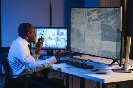 African American security guard looking at desktop computer screen while speaking on walkie talkie.