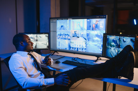 African American security officer or dispatch uses computer and walkie talkie, looks at screens.