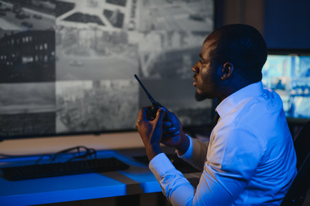 African American security guard looking at desktop computer screen while speaking on walkie talkie.