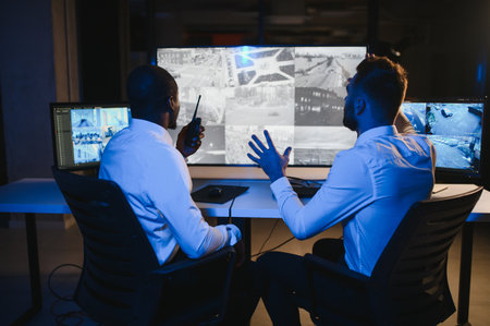 Two security guards sitting by workplace in front of computer monitors and observing cctv video on...