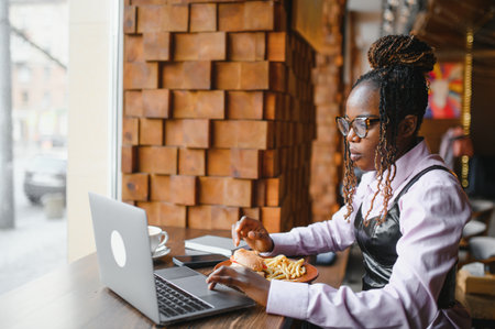 African woman eating fries and burger and working on laptop in cafe.の写真素材