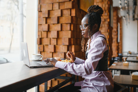 African woman eating fries and burger and working on laptop in cafe.の写真素材