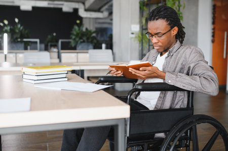 Young man with a disability using a wheelchair, reading a book while focused at his desk in a bright, modern office environmentの写真素材