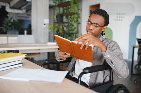 Young african american man using wheelchair reading textbook, focused on studying in bright, accessible office spaceの写真素材