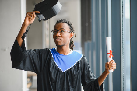 Proud graduate wearing gown, holding his graduation hat and diploma, celebrating successful completion of studiesの写真素材