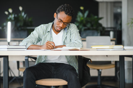 Young man taking notes from textbook, preparing exam at university library, focused student learning and studyingの写真素材