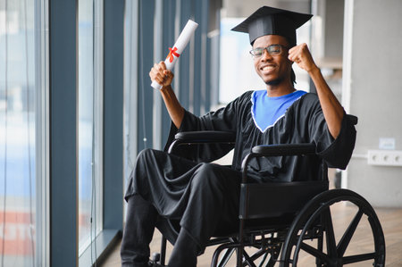 Young black man in graduation gown and mortarboard sitting in wheelchair, holding his diploma and celebrating his academic achievementの写真素材
