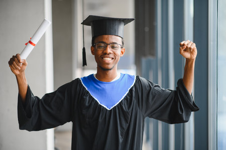 Young man celebrating his graduation with a diploma in hand, smiling and wearing a graduation gown and cap inside university buildingの写真素材