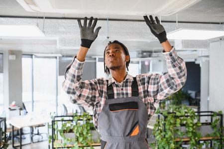 Young african american male technician wearing gloves, repairing air conditioning ventilation on...