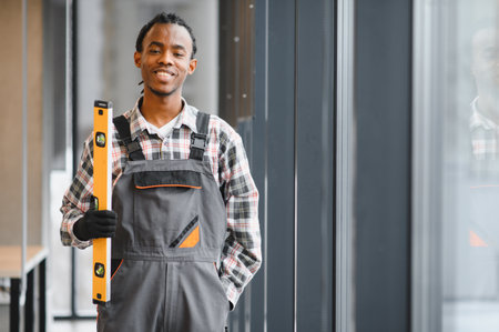 Smiling african american construction worker holding level tool standing by window, showcasing...