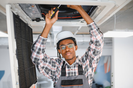 Young african american hvac technician wearing safety hardhat and glasses, using screwdriver while repairing air conditioning ventilation systemの写真素材