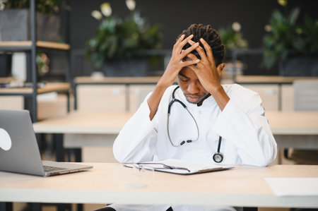 Young male doctor with a stethoscope, sitting at a desk with a laptop, showing stress and fatigue in a modern office environmentの写真素材