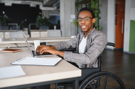 Young man with disability using laptop, working remotely in modern office, representing diversity and inclusion in workplaceの写真素材