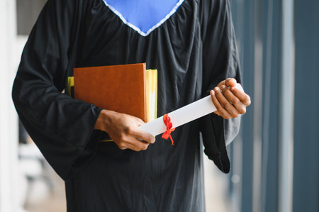 Graduate student proudly holding diploma and books, celebrating academic achievement at university graduation ceremonyの写真素材
