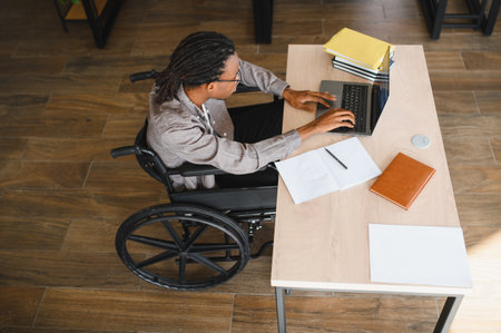 High angle view of a focused young black man with dreadlocks using a laptop at his desk while sitting in a wheelchairの写真素材