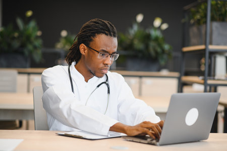 Focused male doctor using laptop, managing patient files and medical records in bright hospital officeの写真素材