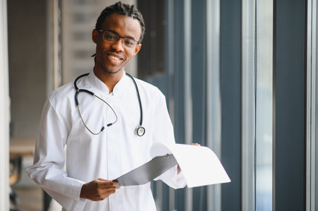African american male doctor reviewing medical documents by a window, wearing a stethoscope and glasses, conveying professionalism and careの写真素材