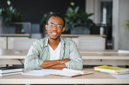 Portrait of a smiling African American male university student sitting at a desk with crossed arms, surrounded by books and notes, in a bright modern library or classroomの写真素材