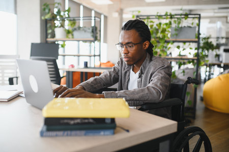 Young man with disability using laptop, concentrating on his online studies or work in a bright, modern office spaceの写真素材