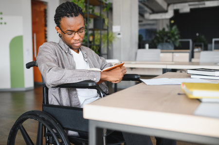 Young black man with disability reading textbook, preparing for exam in accessible college library, embracing inclusive educationの写真素材