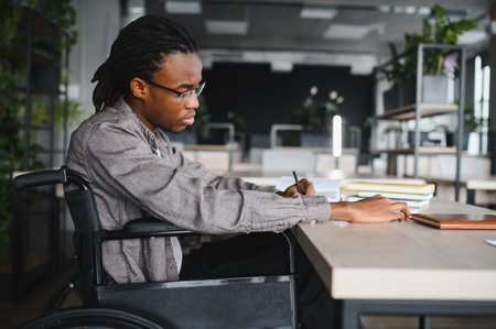 Young man using wheelchair writing notes while studying at desk in modern university library,...