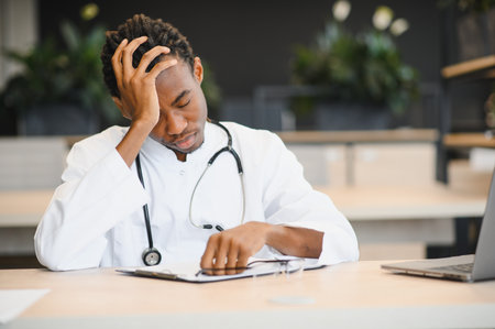 Young african american doctor sitting at desk with head in hand, exhausted after long shift at hospitalの写真素材