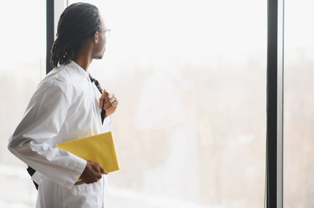 Young black medical student holding books and looking out window, contemplating his future medical careerの写真素材