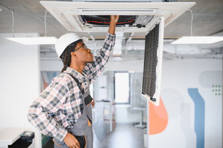 Young african american hvac technician inspecting air conditioning unit on the ceiling wearing hardhat and overallsの写真素材