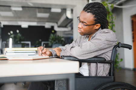 Young black man with disability using a wheelchair, concentrating on his studies and taking notes in a bright, modern office environmentの写真素材