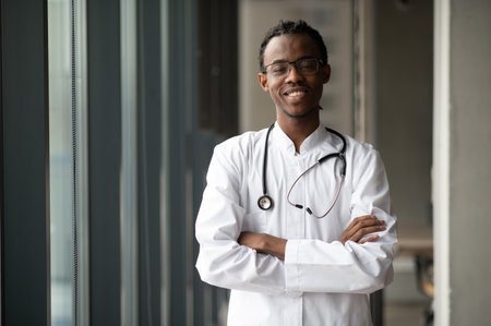 Portrait of a confident African American male doctor smiling with arms crossed, wearing a white coat and stethoscope in a modern hospital corridorの写真素材