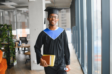 Happy african american graduate student walking in university corridor, wearing graduation gown and holding books and diploma, smilingの写真素材