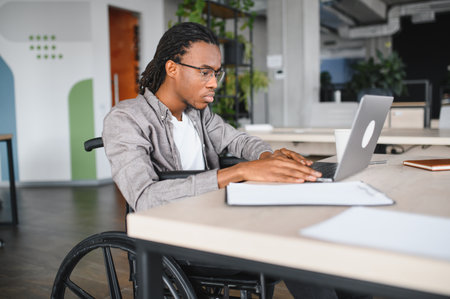 African American student in a wheelchair focused on studying at a laptop in a modern, accessible workspace with greenery and natural lightの写真素材