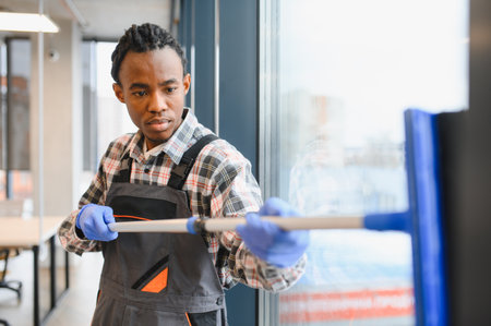 Young african american man wearing blue gloves and gray overalls cleaning windows with squeegee
