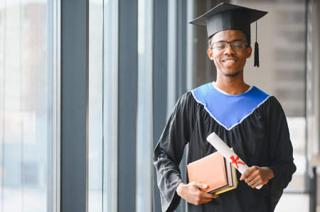 Smiling african american medical student wearing graduation gown holding diploma and books in university corridorの写真素材