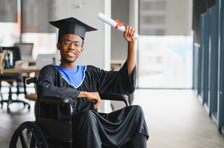 Proud black student in graduation gown and mortarboard, sitting in a wheelchair and holding his diplomaの写真素材