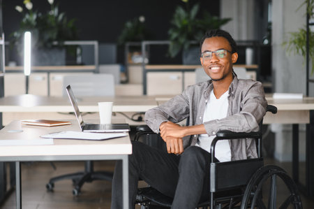 African American student in a wheelchair studying at a desk with a laptop, symbolizing inclusivity and education in a modern workspaceの写真素材