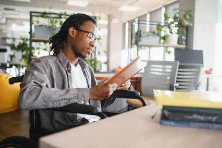 Young black man using wheelchair reading a book while studying in a bright, modern office environmentの写真素材