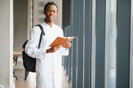 Young african american medical student wearing white coat and backpack, reading book and studying for university exam in corridorの写真素材