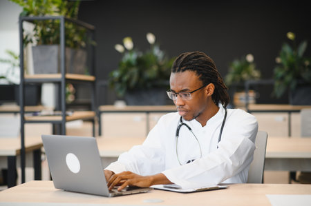 African american male doctor with stethoscope using laptop in a contemporary office setting, surrounded by plants and stylish decorの写真素材