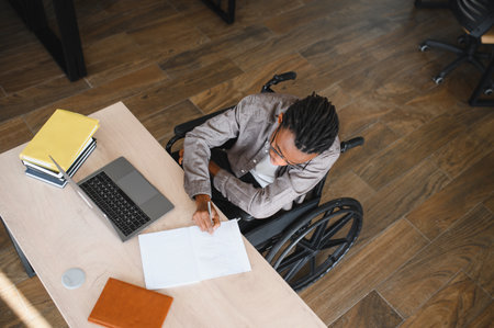 High angle view of a young African American man using a wheelchair, diligently taking notes at a desk in a contemporary office space, showing accessibility and inclusionの写真素材