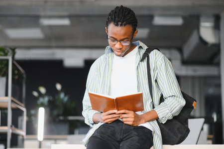 Young black man studying in a university library, deeply focused on reading a textbook and absorbing...