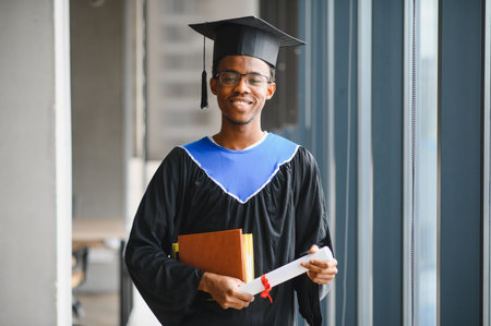 Smiling african american graduate student wearing graduation gown and cap holding diploma and books in university corridorの写真素材
