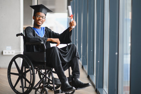 Young man in graduation gown and mortarboard sitting in wheelchair and showing his graduation...